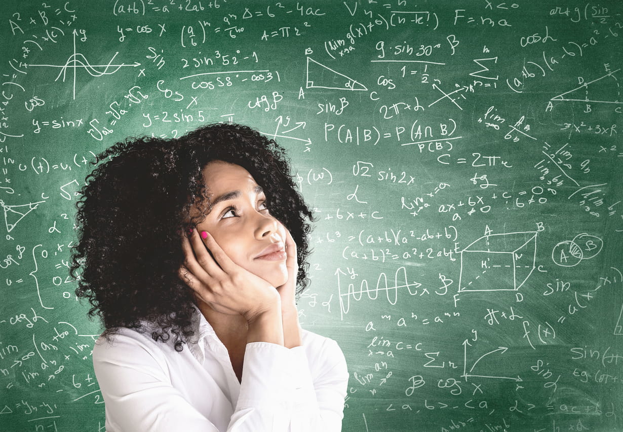 Smiling young African American woman in white shirt looking at blackboard with formulas. Concept of education and science.