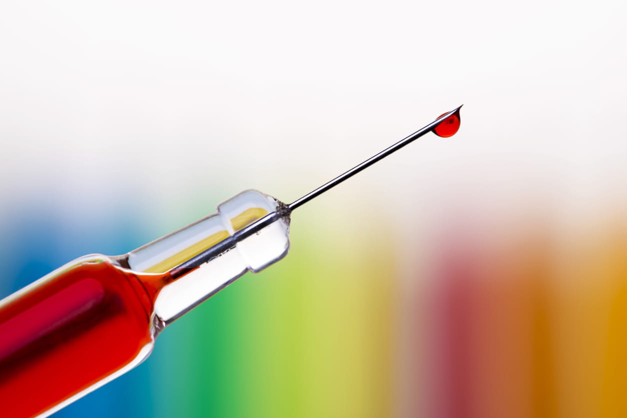 Macro of a single drop of blood hanging at the end of a syringe needle over blurred rainbow background.