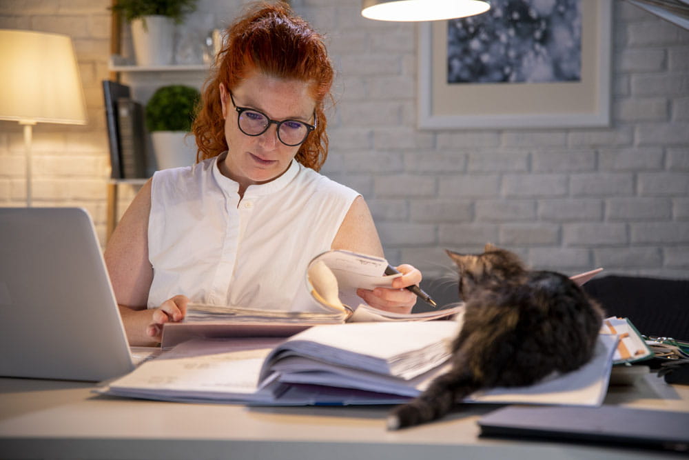Stressed businesswoman with glasses sitting behind home office desk, cat is watching her.